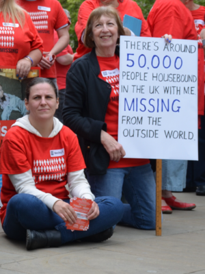 Photo of two women wearing Missing Millions T-shirts at a protest. One is a young woman sat on the concrete. The other woman is smiling and holding a sign saying "There's around 50,000 people housebound in the UK with ME and missing from the outside world."