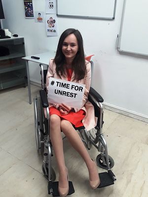 A young girl wearing red and white sits in a manual wheelchair, holding a sign saying #Time for Unrest.